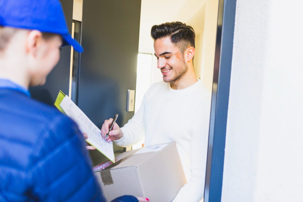 Hombre con barba y camisa blanca firma nota de entrega de pedido, la repartidora viste uniforme de chaqueta y gorra azul